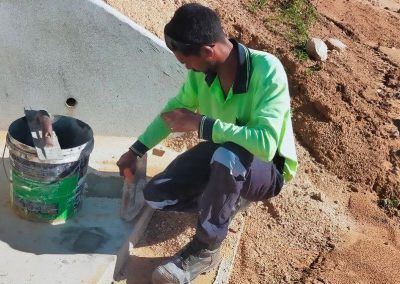 Person in green shirt using a trowel on concrete, next to a bucket. Outdoor setting, dirt and sunlight.