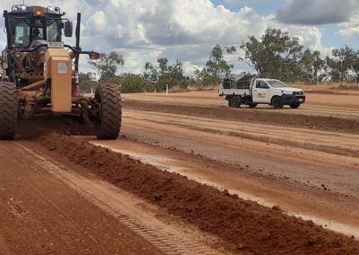 A grader levels a dirt road, with a white truck in the background. The road is red-brown.