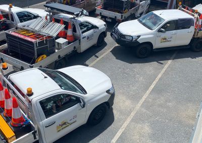 Several white utility trucks parked on asphalt. Cones and equipment visible.