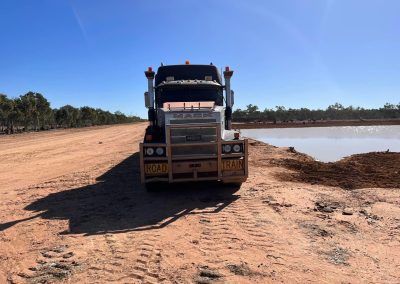Semi-truck parked next to a body of water in a rural, arid landscape; clear, sunny day.