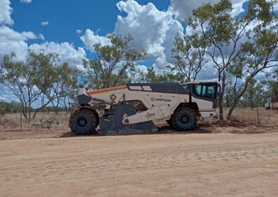 Road milling machine on a dirt road under a blue sky with trees.