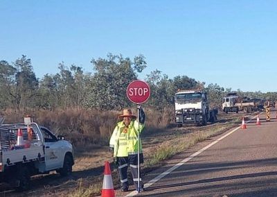Person in safety vest holding stop sign on a road with trucks and cones.