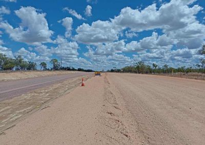 Dirt road and paved road under cloudy blue sky; trees and orange cone are visible.