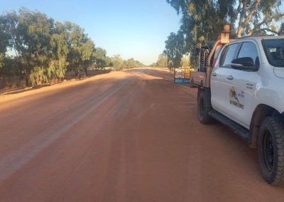 Red dirt road with trees on the sides, white truck parked on the right.