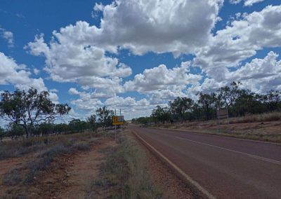Road through a rural landscape with trees, under a blue sky with white clouds.