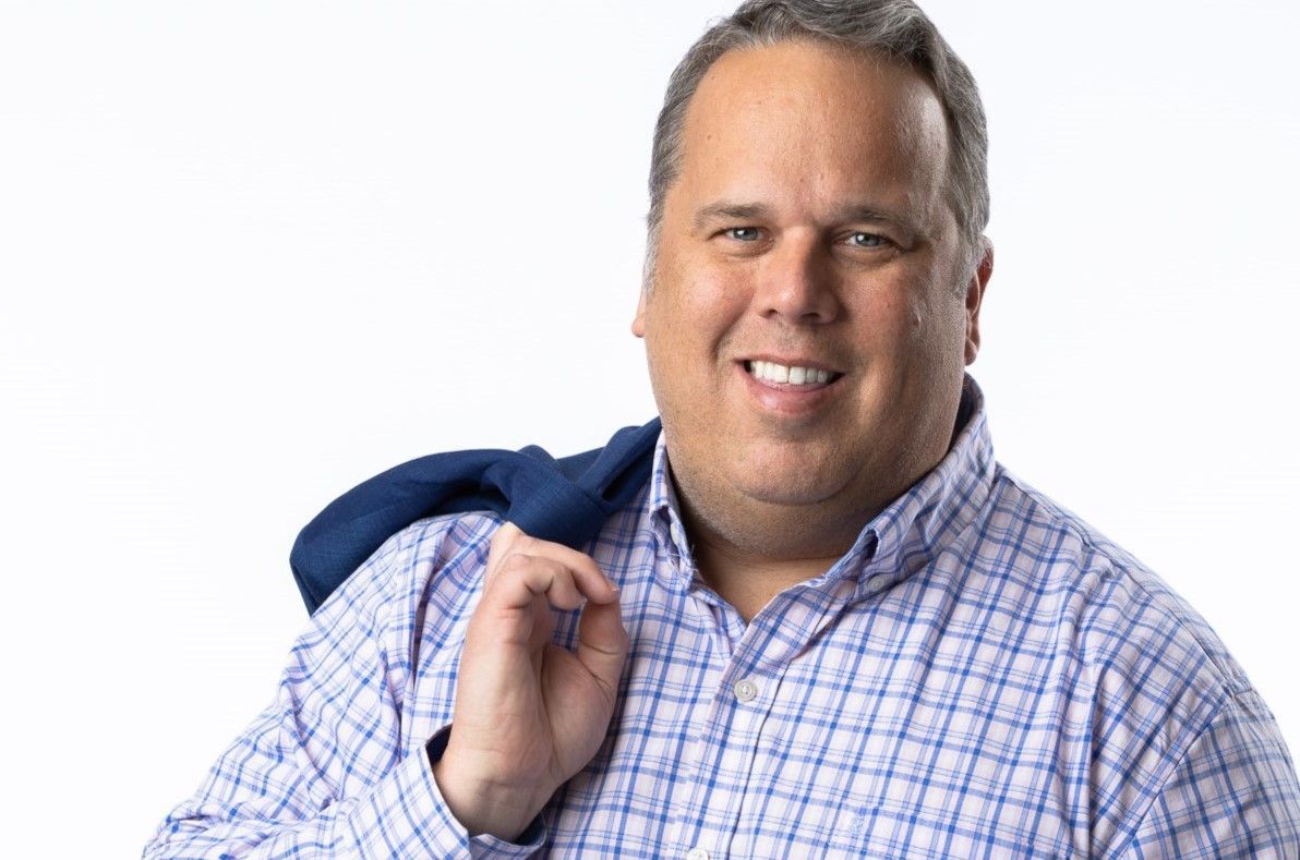 A man in a plaid shirt is smiling and holding a blue jacket over his shoulder.