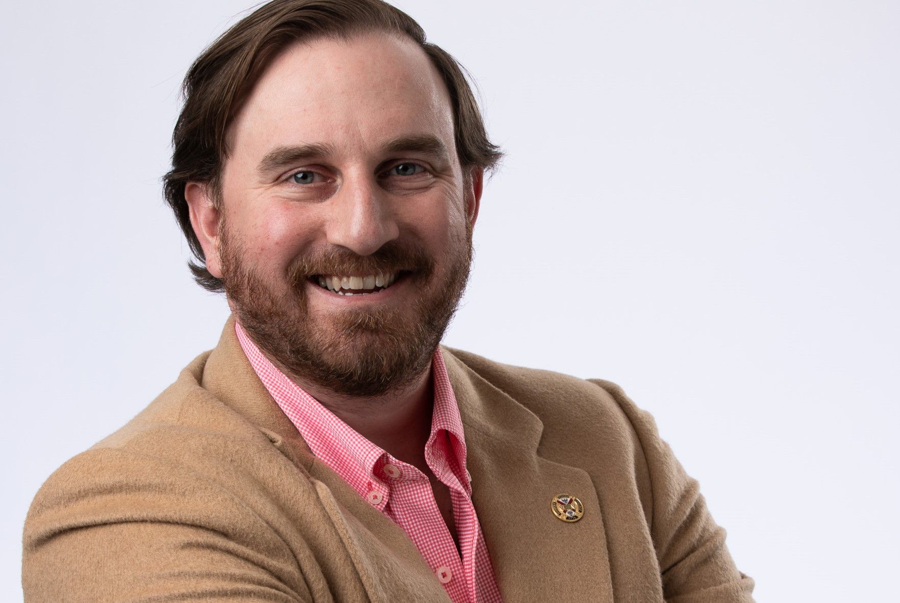 A man with a beard is wearing a tan jacket and a pink shirt.