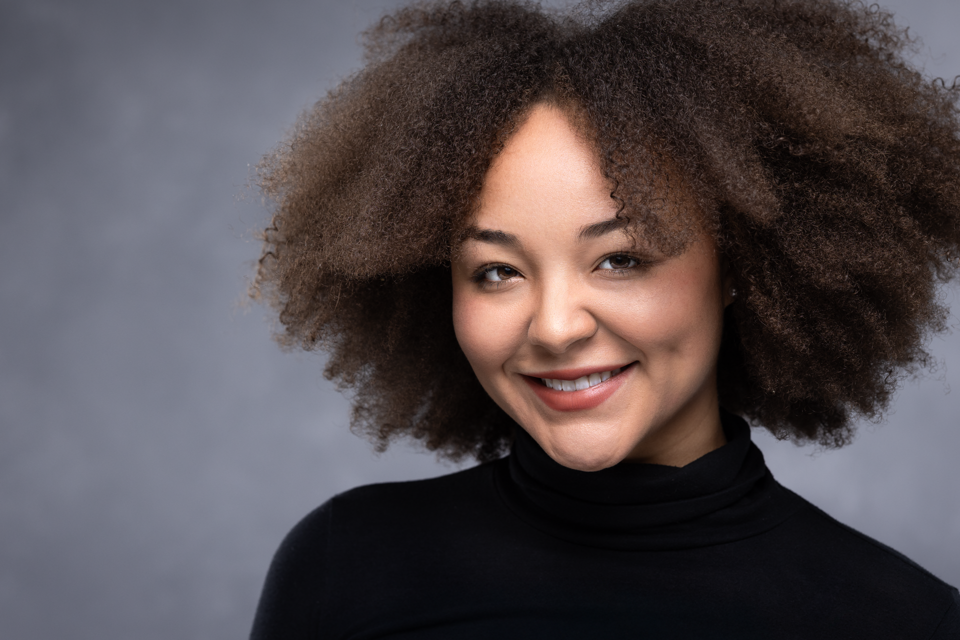 A woman with curly hair is wearing a black turtleneck and smiling.