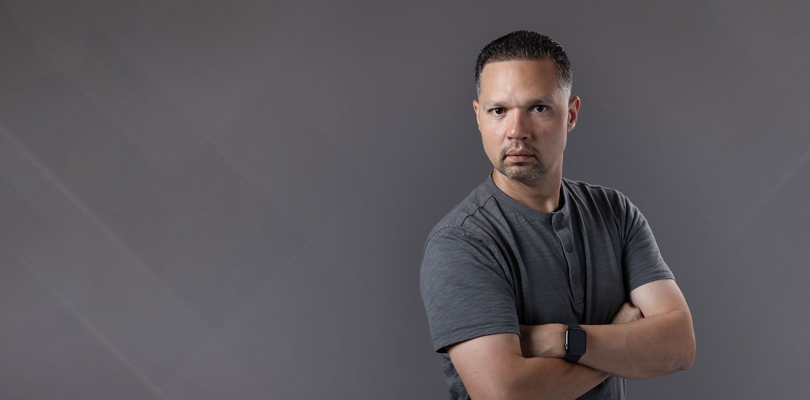 A man in a gray shirt is standing with his arms crossed and looking at the camera.