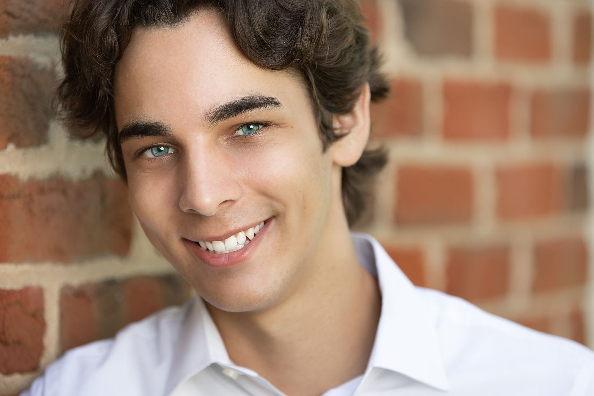 A young man in a white shirt is smiling in front of a brick wall