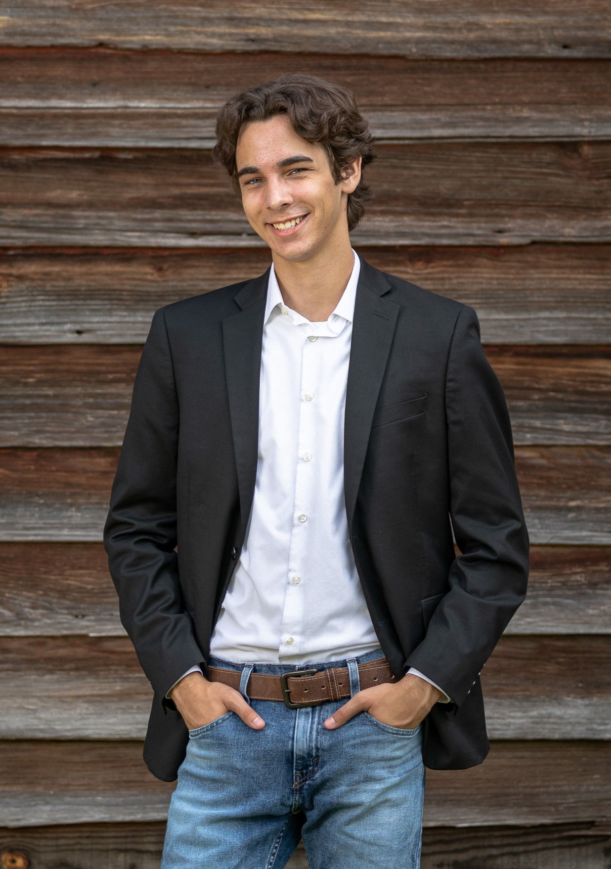 A young man wearing a black jacket and jeans is standing in front of a wooden wall
