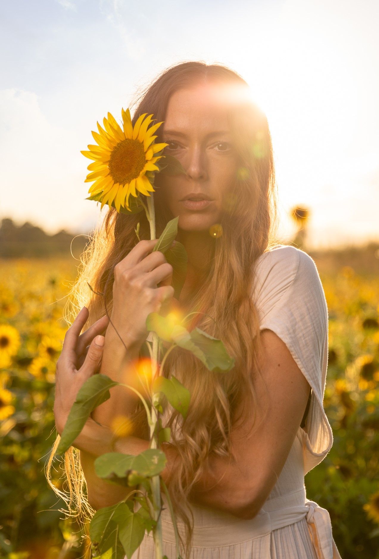 A woman is holding a sunflower in front of her face in a field of sunflowers.
