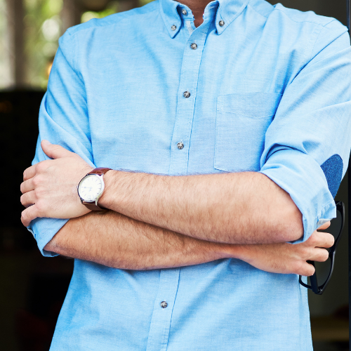 Man with arms crossed, wearing a blue button-down shirt and a watch. Holding glasses.