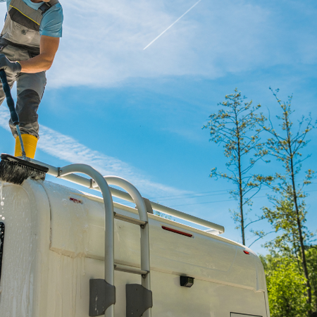 Person washing a white RV roof with a long brush on a sunny day.