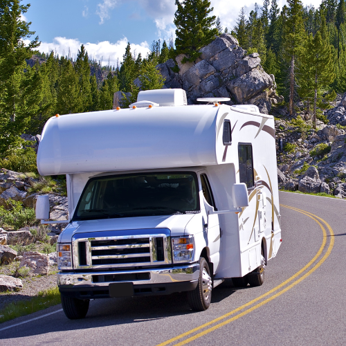 White RV driving on a winding road through a forest with large rock formations.