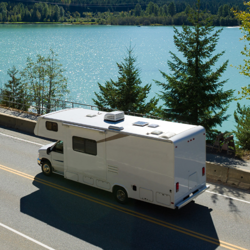 White RV on a road next to a lake with mountains and trees in the background. Sunny day.