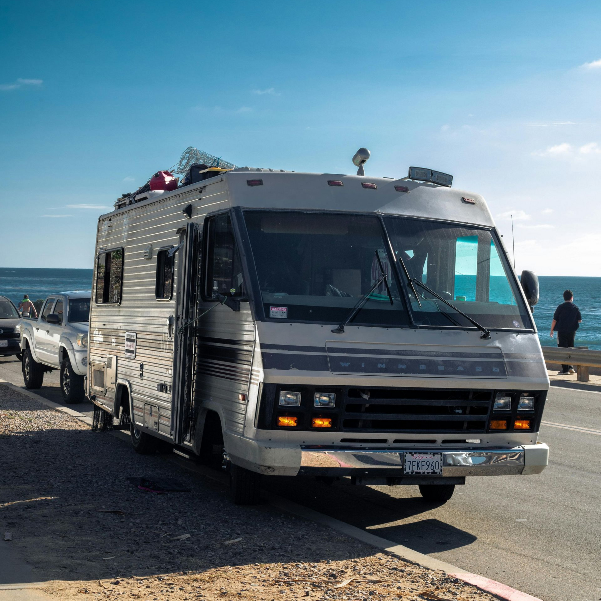 RV parked on a paved road near the ocean under a blue sky; a car and person are also visible.