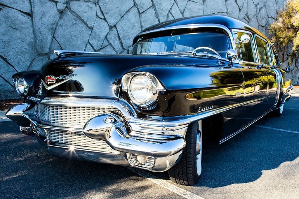 Black vintage Cadillac parked in front of a stone wall, showcasing chrome detailing and a glossy finish.
