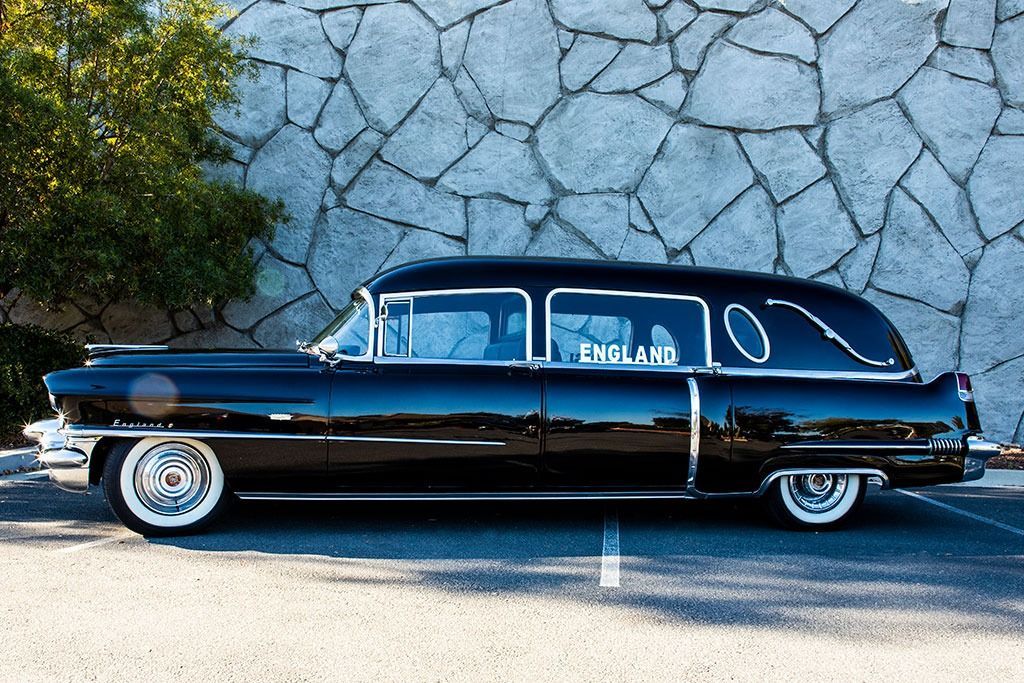 Black hearse parked in front of a stone wall; sunny day.