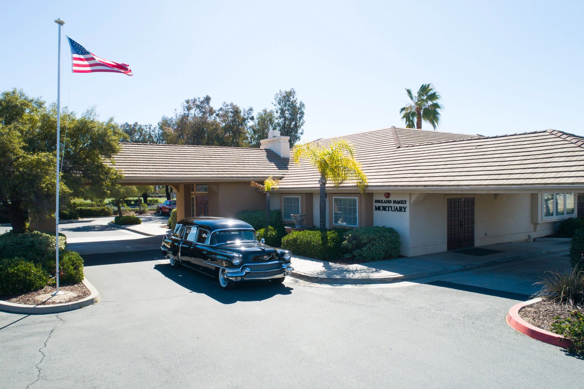A black hearse parked in front of a beige building with a US flag on a sunny day.