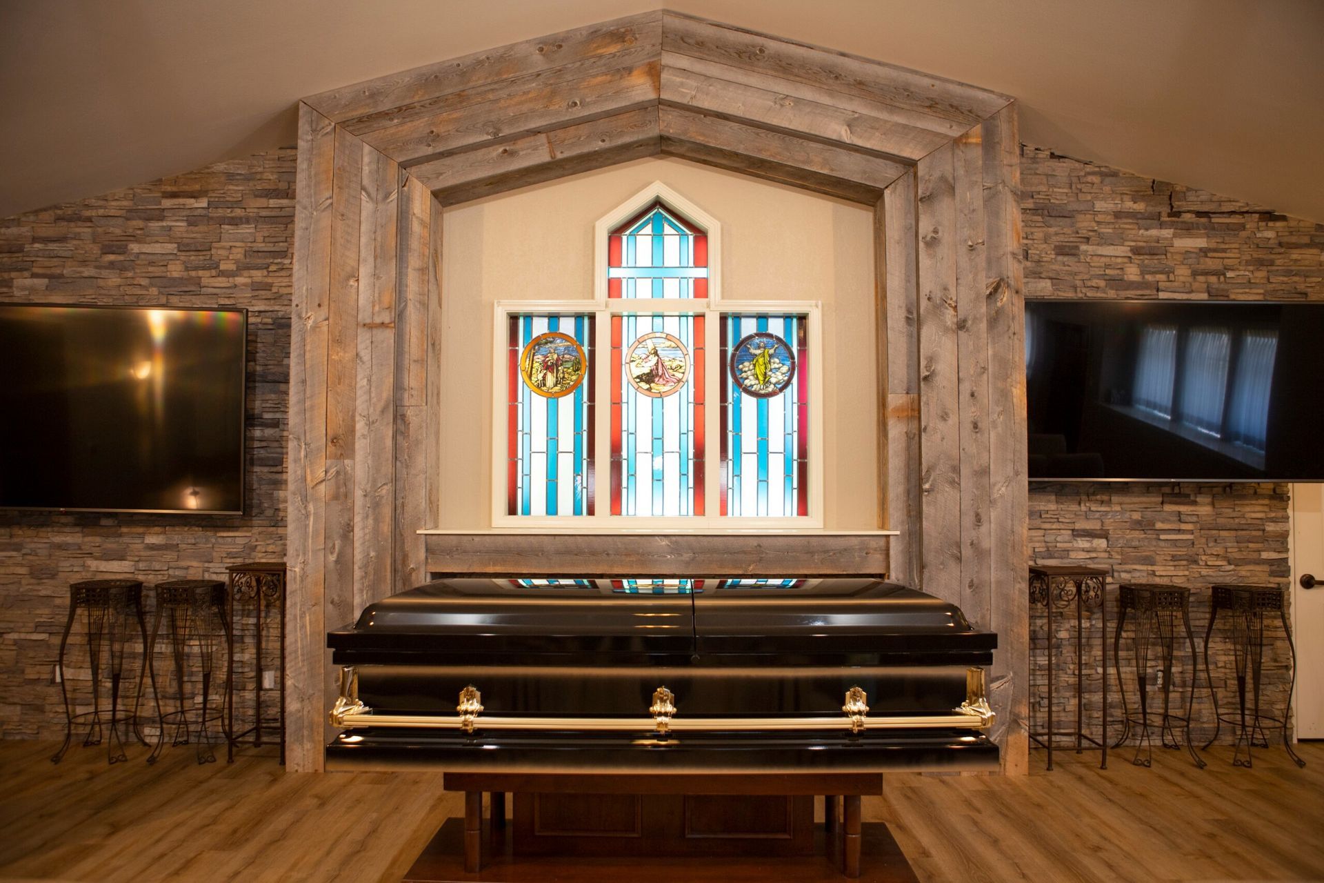 A black casket on a wooden stand in a funeral home, with TVs and stained glass.