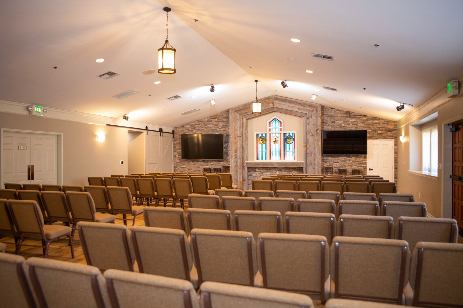 Rows of beige chairs face a sanctuary with a stained glass window, two screens, and brick accents.