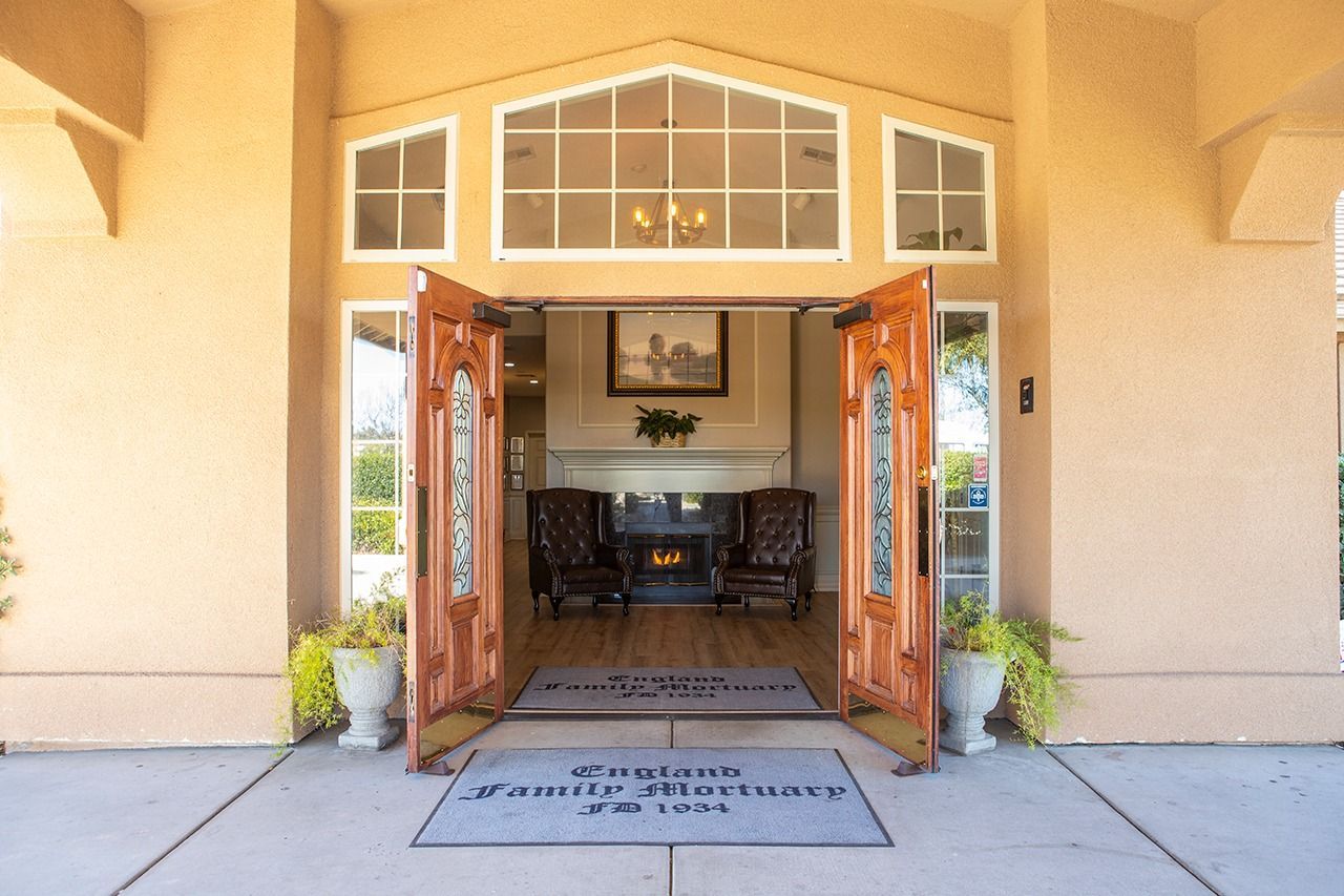 Beige building entrance with open wooden doors, welcome mats, and potted plants.