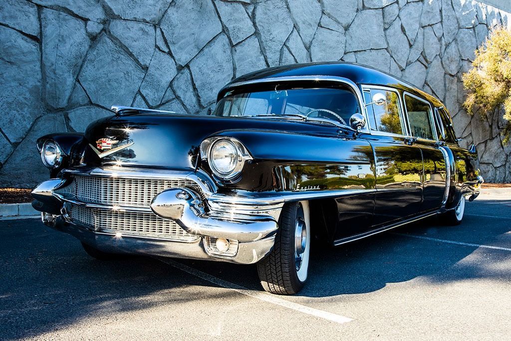 Black vintage Cadillac on asphalt parking area in front of a stone wall.