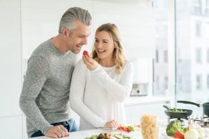 A man is feeding a woman a tomato in a kitchen.