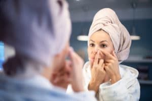 A woman with a towel wrapped around her head is looking at her face in the mirror.