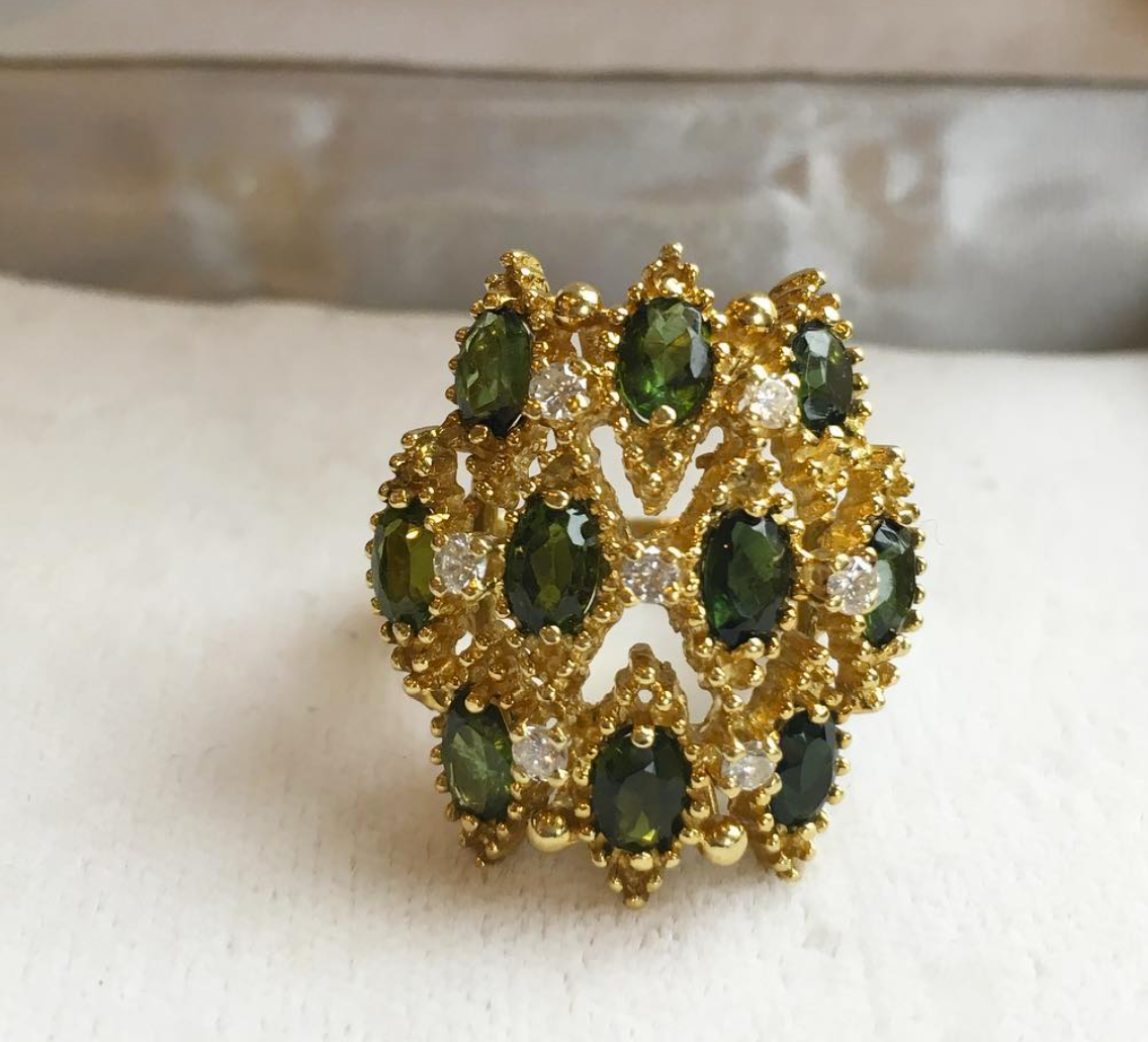 Gold ring with green gemstone clusters in a floral lattice design on a light fabric background