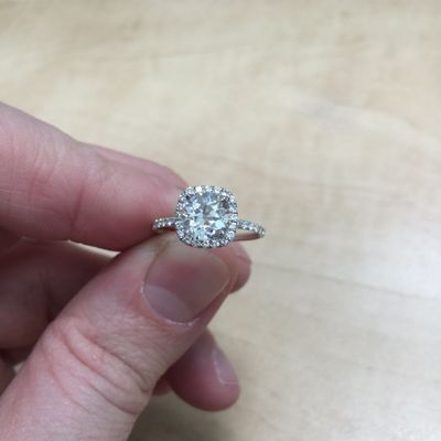 A hand holding a diamond engagement ring with a square halo setting on a light background