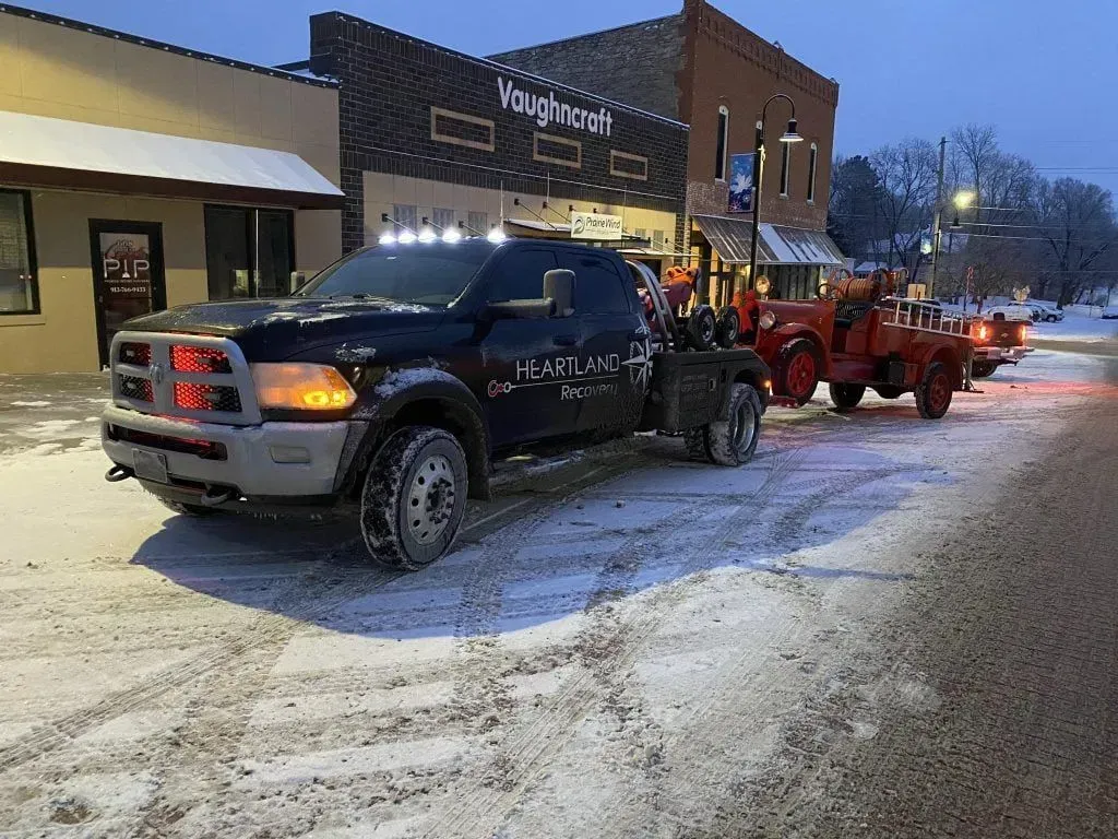 A black flatbed tow truck pulling a red vintage fire truck parked on a snowy, small-town street at dusk.
