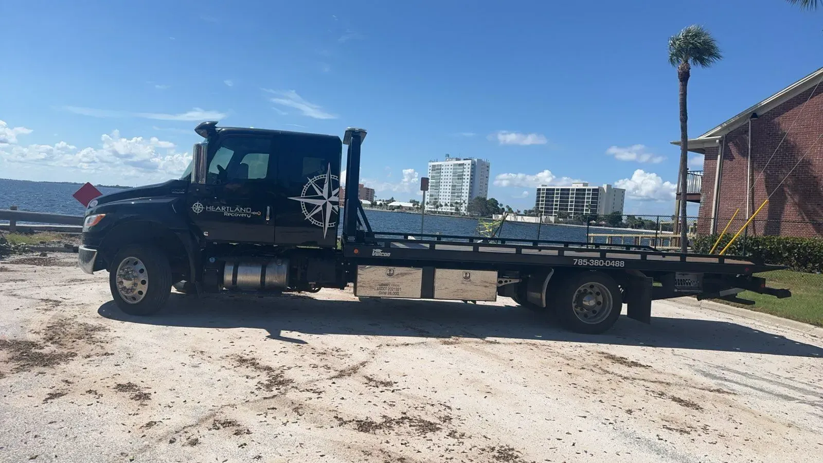 A black flatbed tow truck parked on a gravel surface outdoors with buildings in the background under a blue sky.