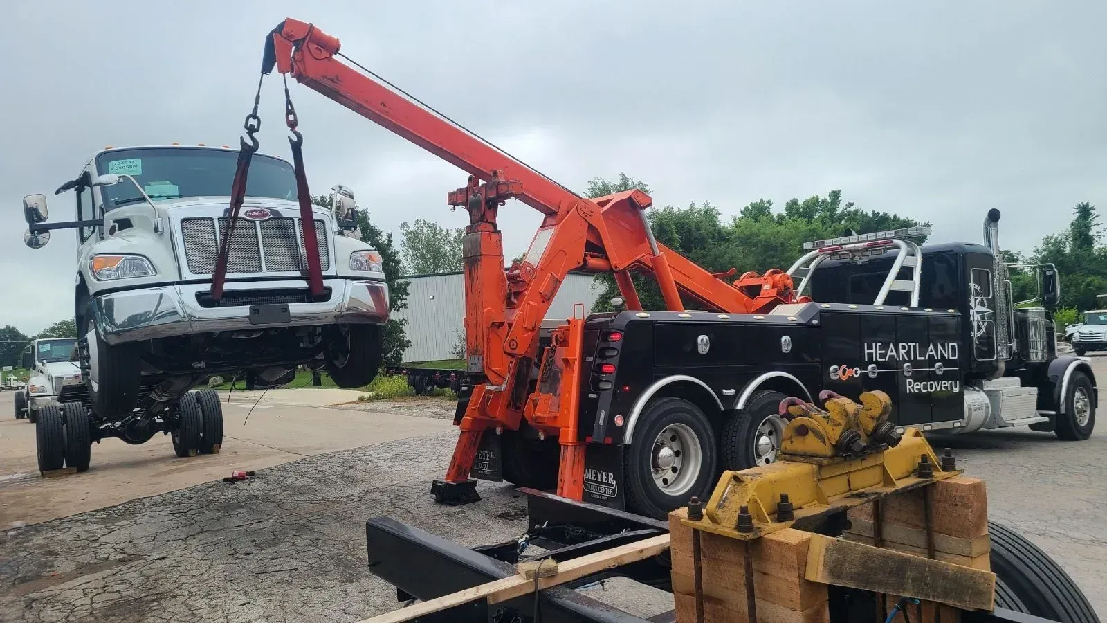 A bright orange tow truck lifts the front of a white semi-truck on a gravel lot under a cloudy sky.
