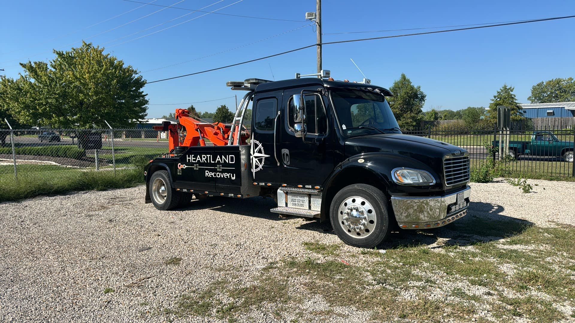 A black tow truck with orange equipment is parked on a gravel lot under a clear blue sky.