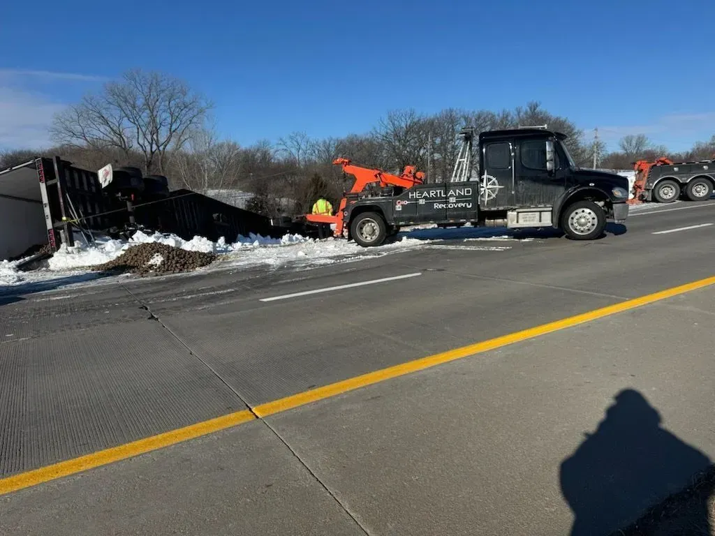 A large orange tow truck connected to an overturned semi-truck trailer on a snowy roadside under a clear blue sky.