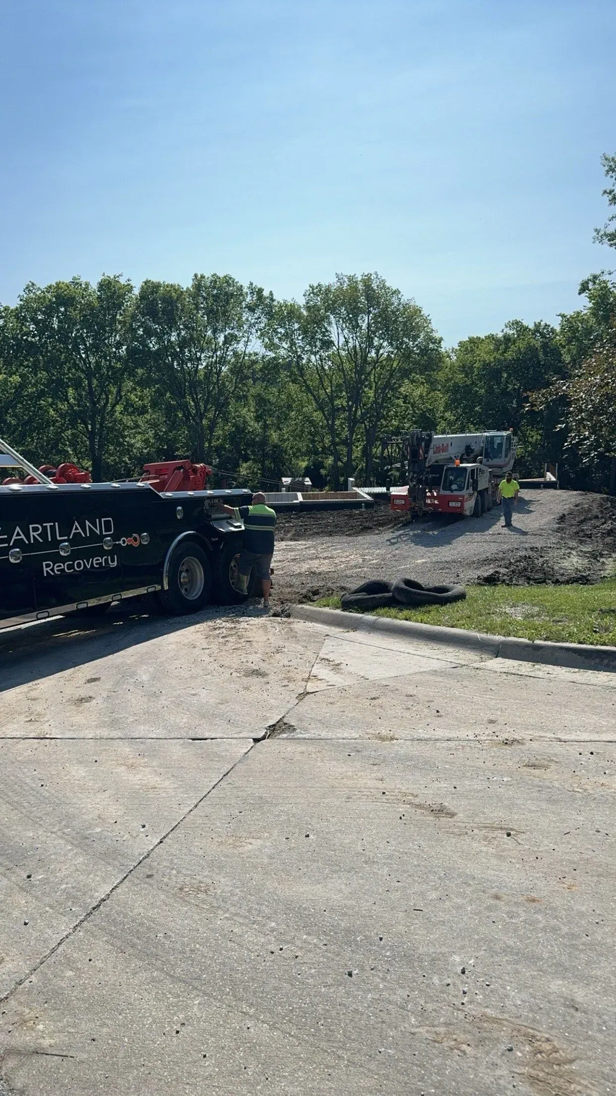 A black truck with text on its side parked on an asphalt lot near a green, wooded area and a light-colored vehicle.