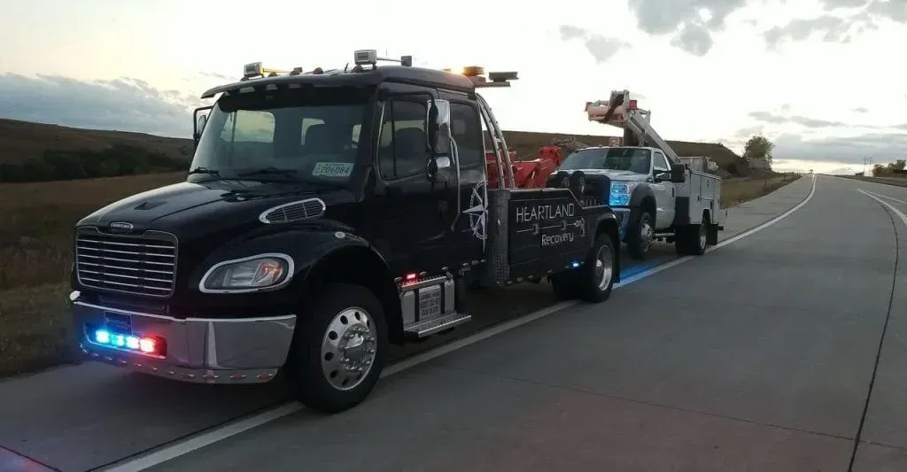 A black tow truck with flashing blue and red lights tows a white utility vehicle along a paved road at dusk.
