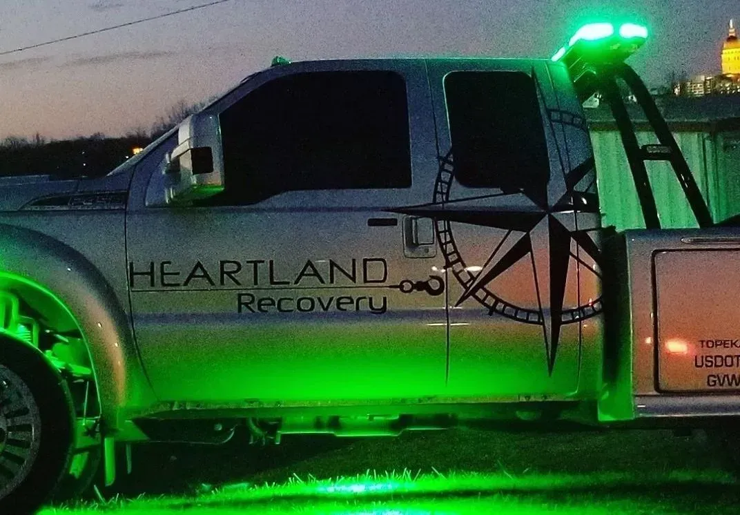 A silver Heartland Recovery tow truck parked at twilight, illuminated by bright green underglow and rooftop lights.