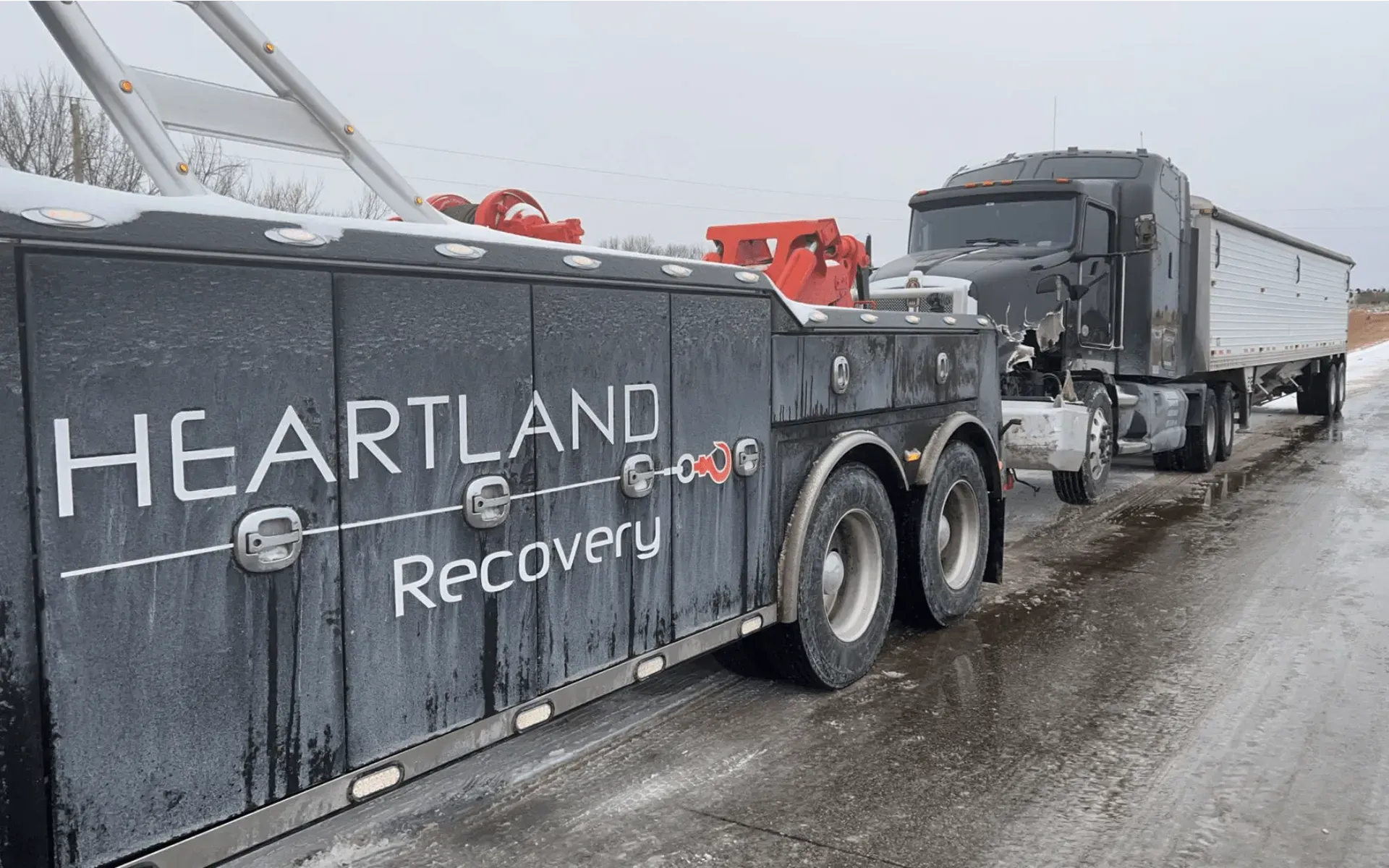A black Heartland Recovery tow truck pulling a semi-truck along a slushy, snow-covered road.