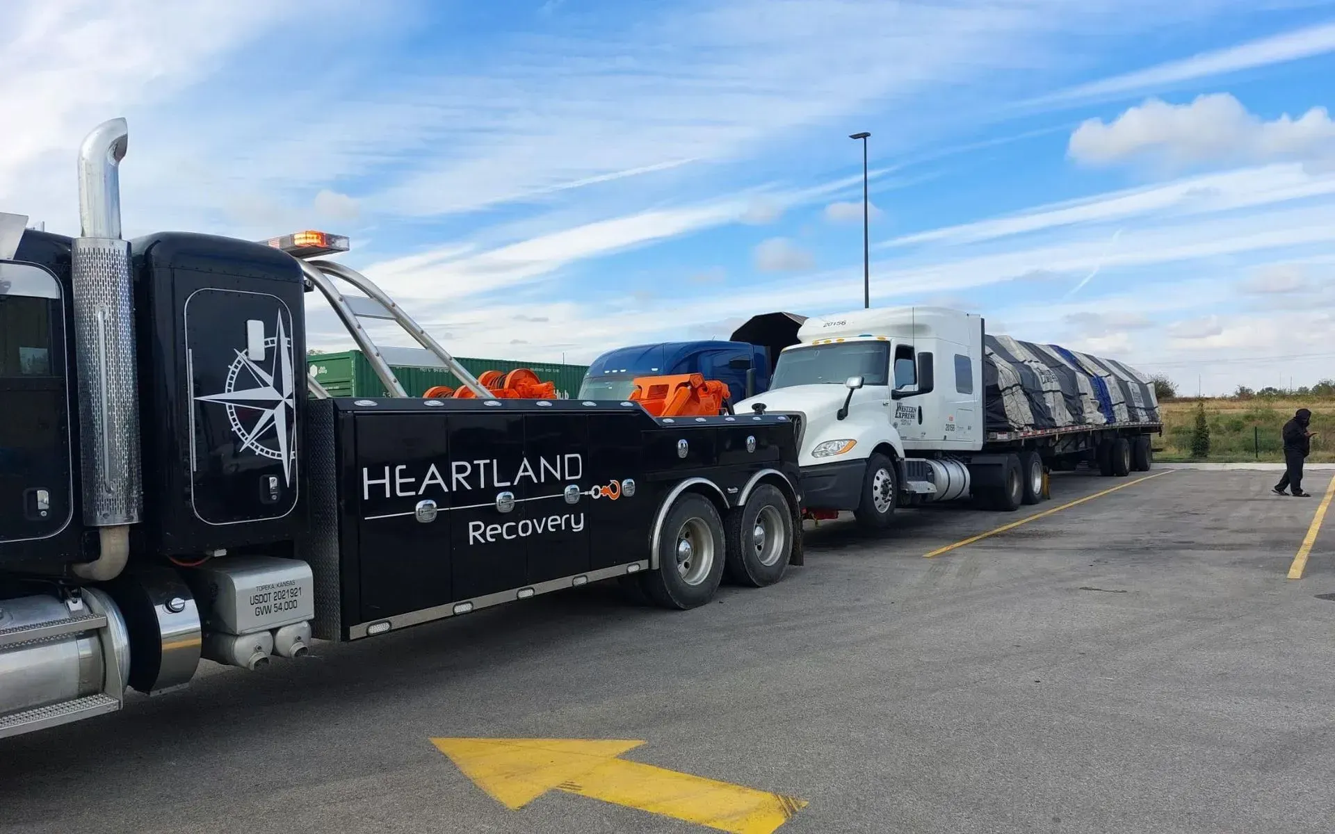 A black Heartland Recovery tow truck towing a white semi-truck with a flatbed trailer in a parking lot.