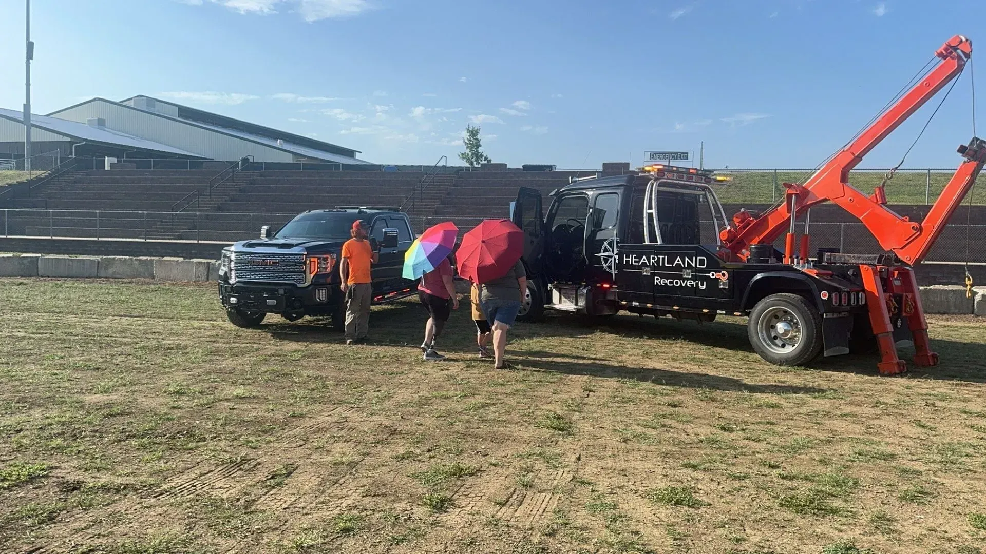 Two people under umbrellas stand between a truck and a wrecker in a grassy field near a large stadium structure.