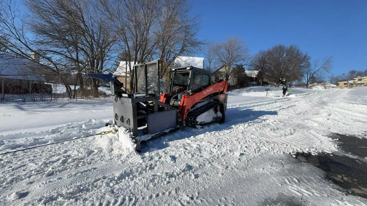 An orange skid steer loader with a snow blower attachment clears a snow-covered residential street on a sunny day.