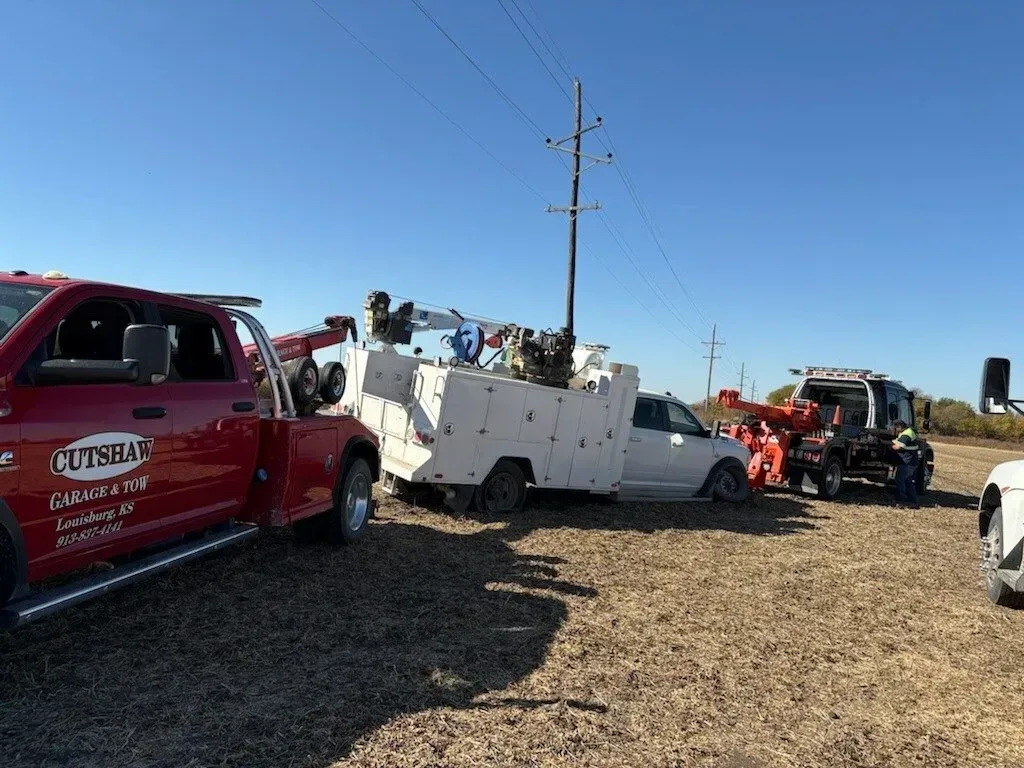 A red tow truck is attached to a white service truck stuck in a field next to a utility pole and a piece of farm equipment.