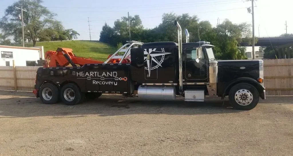 A black tow truck with orange equipment, parked on a gravel lot in front of a wooden fence under a bright sky.