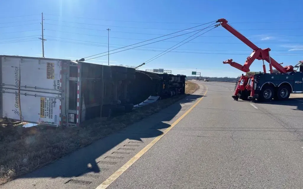 A large red tow truck uses cables to lift an overturned tractor-trailer on the side of a highway under a clear blue sky.