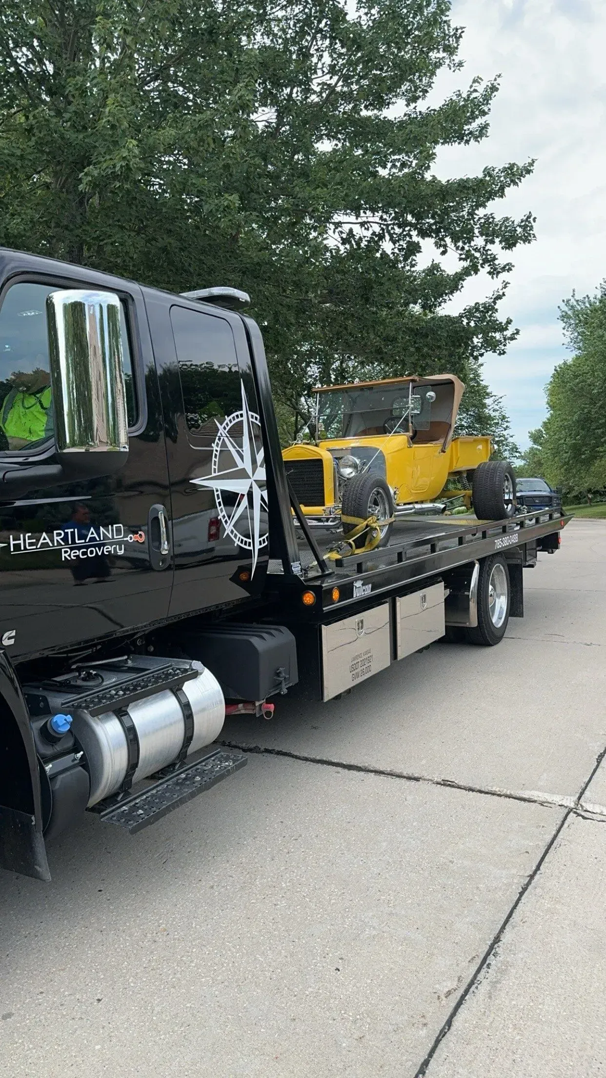 A black flatbed tow truck carrying a small, yellow vintage hot rod on a paved road with trees in the background.