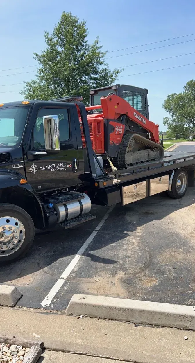 A black flatbed tow truck parked in an outdoor lot carrying a bright orange Bobcat compact track loader on its bed.