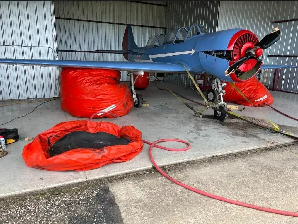 A blue vintage airplane sits inside a hangar, lifted by bright orange pneumatic lifting bags under its wings and fuselage.